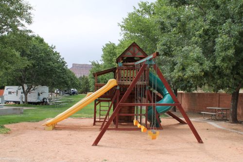 A wooden playground with a slide, swings, and climbing structure, surrounded by trees and picnic tables.