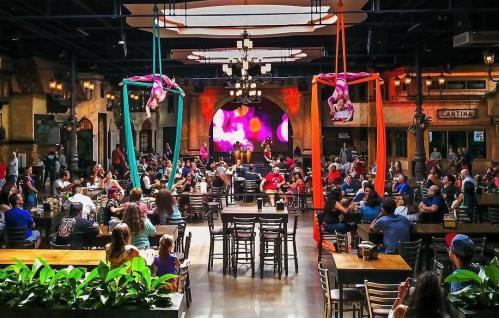 A lively restaurant interior filled with people, featuring aerial performers and colorful decor.