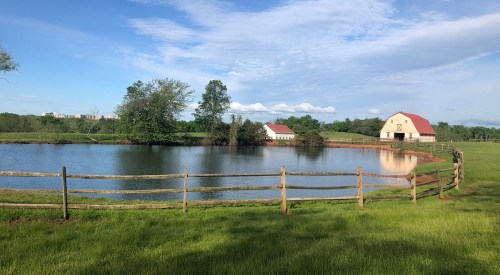 A serene landscape featuring a pond, green grass, and a red-roofed barn surrounded by trees and a wooden fence.
