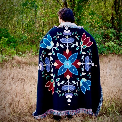A person stands in a field, wearing a colorful, floral-patterned blanket with fringes, surrounded by trees.