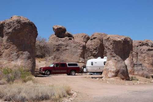 A red truck parked next to a white trailer surrounded by large rock formations under a clear blue sky.