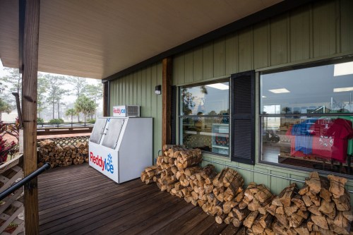 A wooden deck with stacked firewood and a cooler labeled "Ready Ice" next to a shop window displaying colorful shirts.