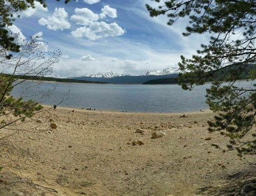 A serene lake surrounded by mountains, with a sandy shore and scattered rocks under a blue sky with clouds.