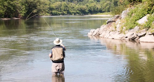 A person fishing in a river, wearing a hat and waders, with trees and rocks lining the shore.