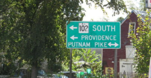 Green road sign indicating directions to South, Providence, and Putnam Pike. Surrounded by trees and buildings.