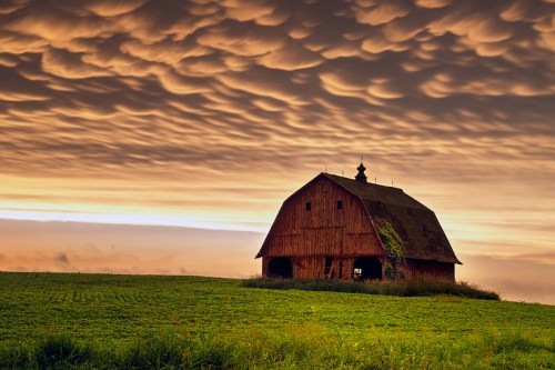 A rustic red barn stands in a green field under a dramatic, cloud-filled sunset sky.