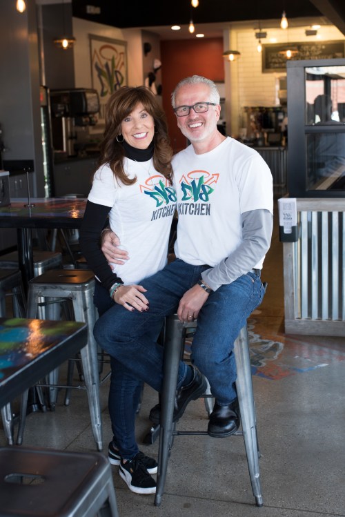 A smiling couple in matching t-shirts sits on bar stools in a cozy kitchen-themed café.