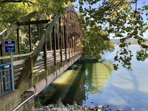 A metal bridge spans a calm river, surrounded by trees, with a "No Fishing" sign nearby.