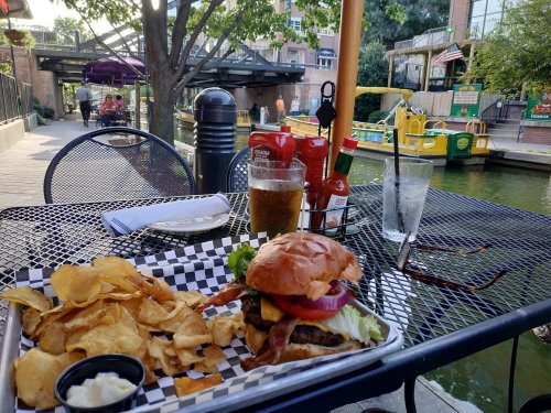 A burger with bacon, lettuce, and tomato, served with chips, a drink, and condiments on a patio by a canal.