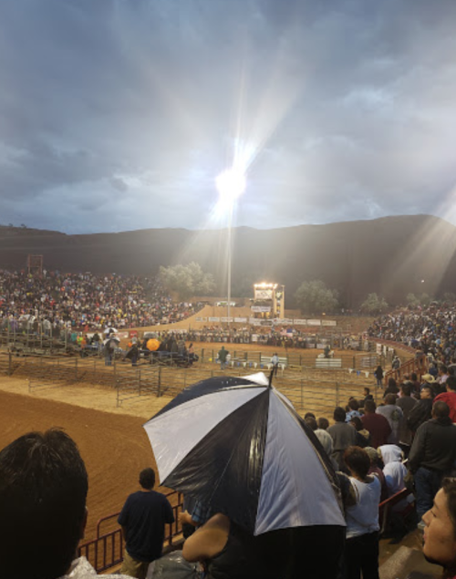 A crowded outdoor arena under cloudy skies, with bright lights illuminating a rodeo event. People are holding umbrellas.