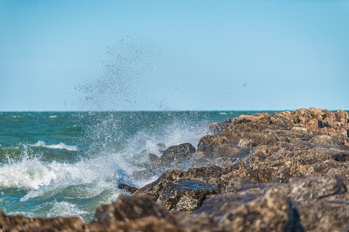 Waves crashing against rocky shoreline under a clear blue sky, creating a spray of water.