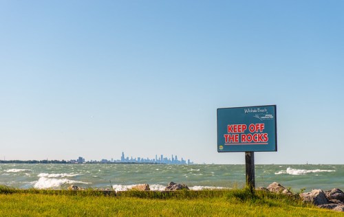 A sign reading "KEEP OFF THE ROCKS" near a beach, with a city skyline visible in the background and waves crashing.