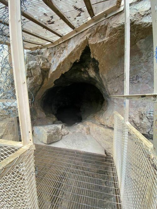 A rocky cave entrance with a metal grate and fencing, surrounded by rough stone walls.