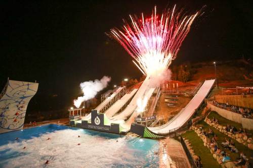 Fireworks illuminate the night sky above a water park with slides and a wave pool, as spectators enjoy the show.