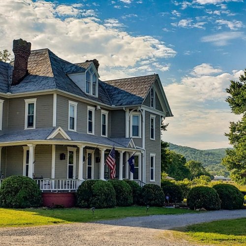 A charming Victorian-style house with flags, surrounded by lush greenery and a scenic mountain backdrop under a blue sky.