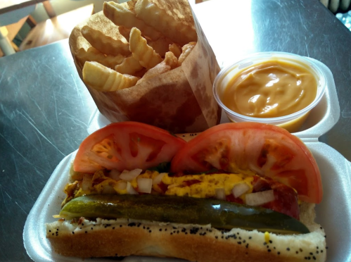 A hot dog topped with tomatoes, pickles, and onions, served with crinkle-cut fries and cheese sauce in a cup.
