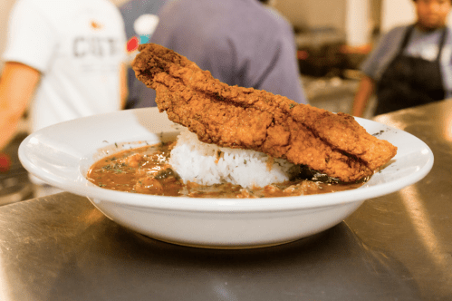 A plate of fried fish atop a mound of rice, served with a flavorful sauce and garnished with herbs.