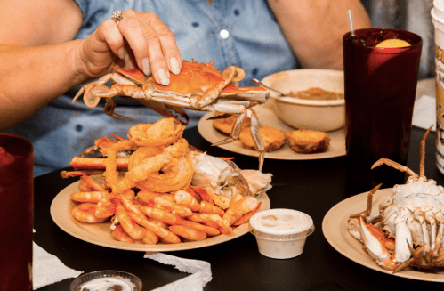A hand lifts a crab from a plate piled with shrimp, onion rings, and a bowl of soup, with drinks in the background.