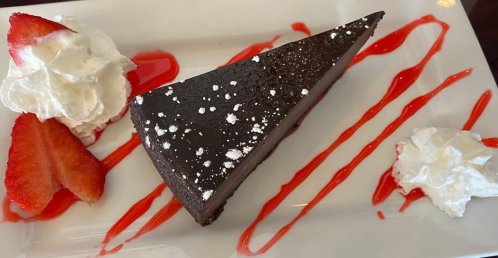 A slice of chocolate cake on a plate, garnished with whipped cream, strawberries, and a drizzle of red sauce.