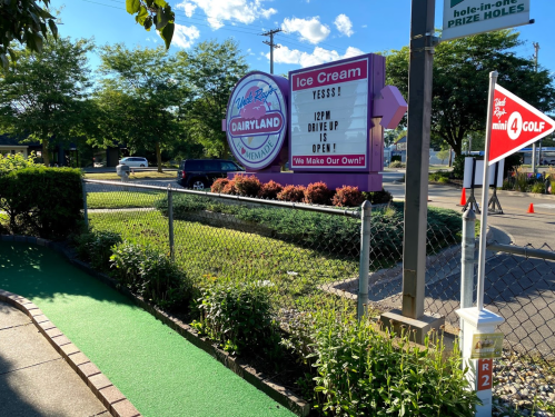 Sign for Dairyland Ice Cream with a message about drive-up hours, surrounded by greenery and a mini-golf area.