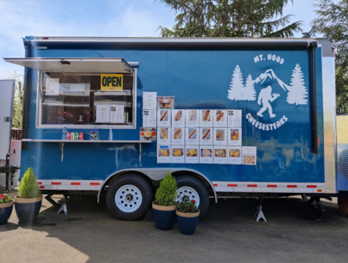 A blue food truck with "Mt. Hood Chefs" logo, open sign, and a menu displayed on the side, surrounded by potted plants.