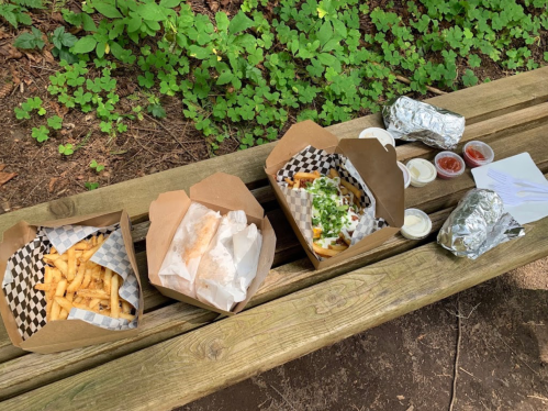 Three takeout boxes of food, including fries and a wrapped burrito, placed on a wooden bench in a green outdoor setting.
