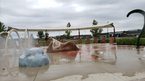A splash pad with colorful water features and seating under a canopy, set against a cloudy sky.