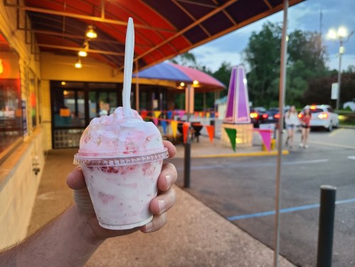 A hand holds a cup of strawberry swirl ice cream outside a colorful ice cream shop.