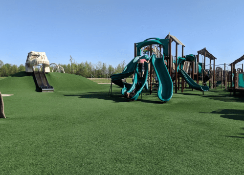 A playground with green turf, featuring slides, climbing structures, and children playing under a clear blue sky.