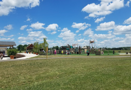A sunny park scene featuring a playground, grassy area, and scattered clouds in a blue sky. Families enjoy the space.
