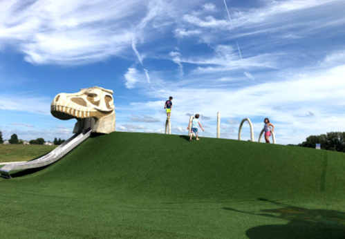 Children playing on a grassy hill with a dinosaur slide under a blue sky with clouds.