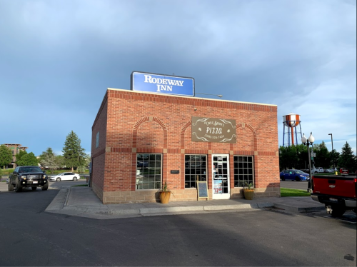 A brick building with a sign for "Rodeway Inn" and a pizza shop, surrounded by parked cars and trees.