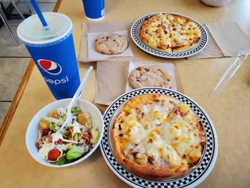 A table with two pizzas, a salad, cookies, and a Pepsi drink on a checkered plate.