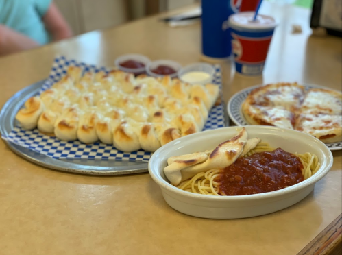 A table with cheesy breadsticks, pizza, and spaghetti with marinara sauce, served with dipping sauces.