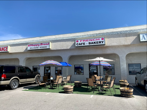 Outdoor seating area with umbrellas in front of a French café and bakery, featuring a sign and nearby parking.