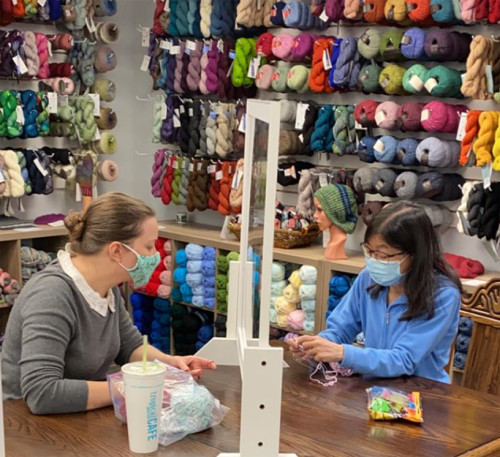 Two women wearing masks sit at a table in a yarn shop, surrounded by colorful yarns, working on a craft project.