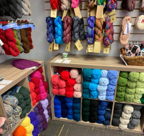 A display of colorful yarns in various shades, organized on shelves and in bins, with a basket of yarn on a table.