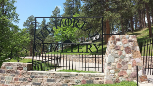 A wrought iron gate with "Memorial Cemetery" inscribed, surrounded by trees and a stone wall.