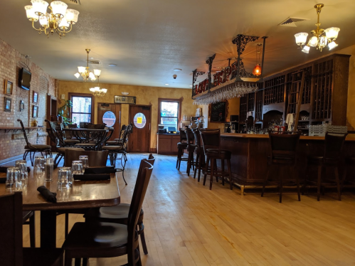 A cozy restaurant interior featuring wooden tables, chairs, and a bar with hanging lights and wine glasses.