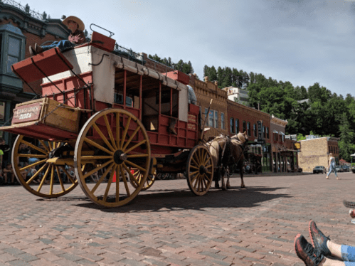 A horse-drawn wagon with a driver sits on a brick street, surrounded by historic buildings and spectators.