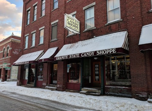 A brick building with a sign reading "Granite State Candy Shoppe," featuring snow-covered awnings and a winter scene.