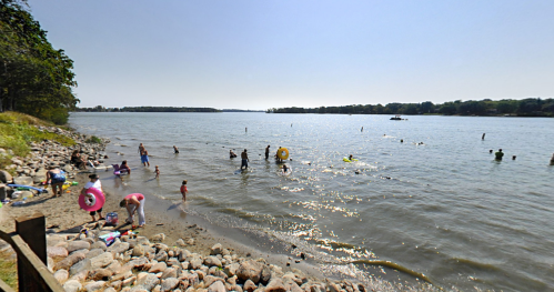 People enjoying a sunny day at a lake, swimming and playing near the shore with colorful floats and rocks.
