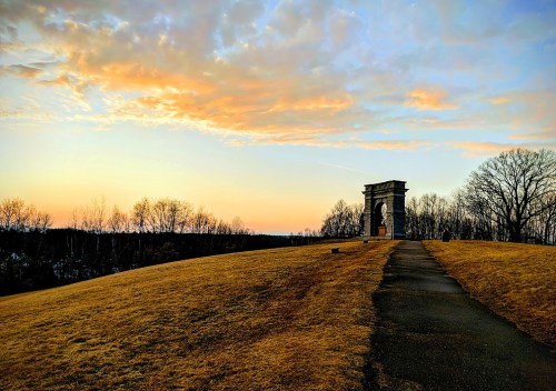 A stone archway on a grassy hill at sunset, with colorful clouds and bare trees in the background.