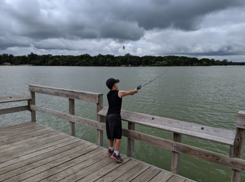 A boy stands on a wooden dock, fishing in a calm lake under a cloudy sky.