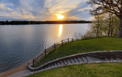 A serene lakeside view at sunset, with a winding staircase leading down to the water and trees lining the shore.