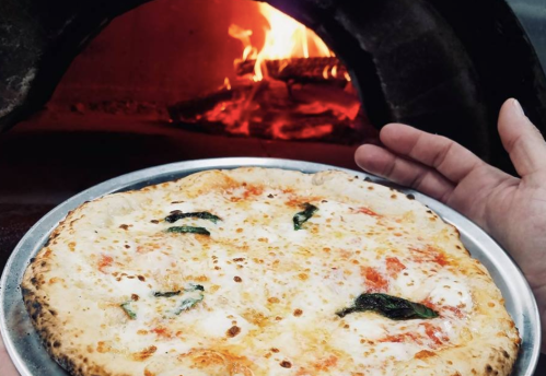 A hand holds a freshly baked pizza with melted cheese and basil, with a wood-fired oven glowing in the background.
