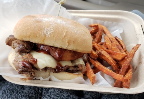 A hearty sandwich with beef, cheese, and BBQ sauce, served with sweet potato fries in a takeout container.