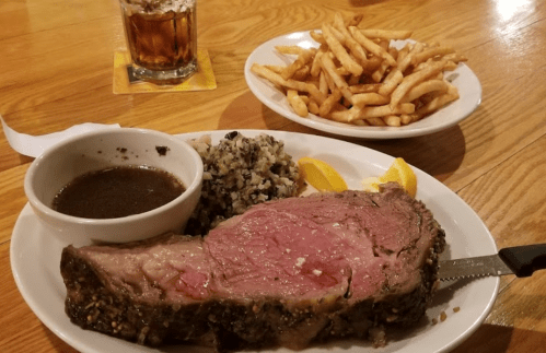 A plate with a large slice of prime rib, gravy, rice, lemon wedges, and a side of fries, with a drink in the background.