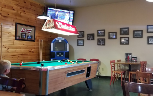 A cozy bar with a pool table, a TV showing a game, and wooden decor, featuring a jukebox and framed photos on the walls.