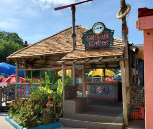 A tropical-themed hut with a thatched roof, labeled "Gilligan's Gully," surrounded by colorful umbrellas and greenery.